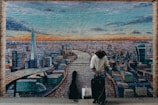 A man in a wide-brimmed hat and textured sweater stands in front of a mural depicting a cityscape. The mural shows a river running through a city, with prominent skyscrapers and an orange and blue sky above. The man has a suitcase and a shopping bag, and a guitar case is visible beside him.