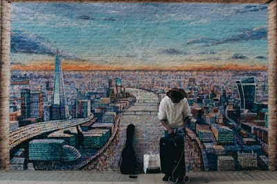 A man in a wide-brimmed hat and textured sweater stands in front of a mural depicting a cityscape. The mural shows a river running through a city, with prominent skyscrapers and an orange and blue sky above. The man has a suitcase and a shopping bag, and a guitar case is visible beside him.