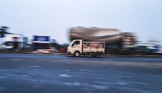A small truck is moving quickly on a road, with motion blur indicating speed. In the background, there are structures and another blurry vehicle, suggesting a busy roadway or urban environment.