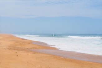 A serene beach scene with golden sand stretching along the coastline. Gentle waves are lapping onto the shore under a bright blue sky with a few scattered clouds.