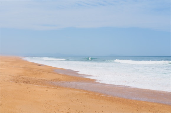 A serene beach scene with golden sand stretching along the coastline. Gentle waves are lapping onto the shore under a bright blue sky with a few scattered clouds.