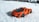 A small, detailed toy sports car with an orange body and black accents is positioned on a snowy surface. The bright color of the car contrasts sharply with the white background, suggesting playfulness and vibrancy.