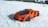 A small, detailed toy sports car with an orange body and black accents is positioned on a snowy surface. The bright color of the car contrasts sharply with the white background, suggesting playfulness and vibrancy.