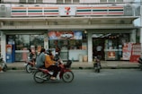 A convenience store with a recognizable red, orange, and green striped logo is situated on a busy street. The storefront is adorned with advertisements and an ATM is located to the left. Several motorbikes are parked in front, and a few individuals wearing helmets are seen riding a motorbike past the store.