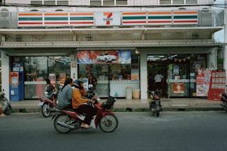 A convenience store with a recognizable red, orange, and green striped logo is situated on a busy street. The storefront is adorned with advertisements and an ATM is located to the left. Several motorbikes are parked in front, and a few individuals wearing helmets are seen riding a motorbike past the store.