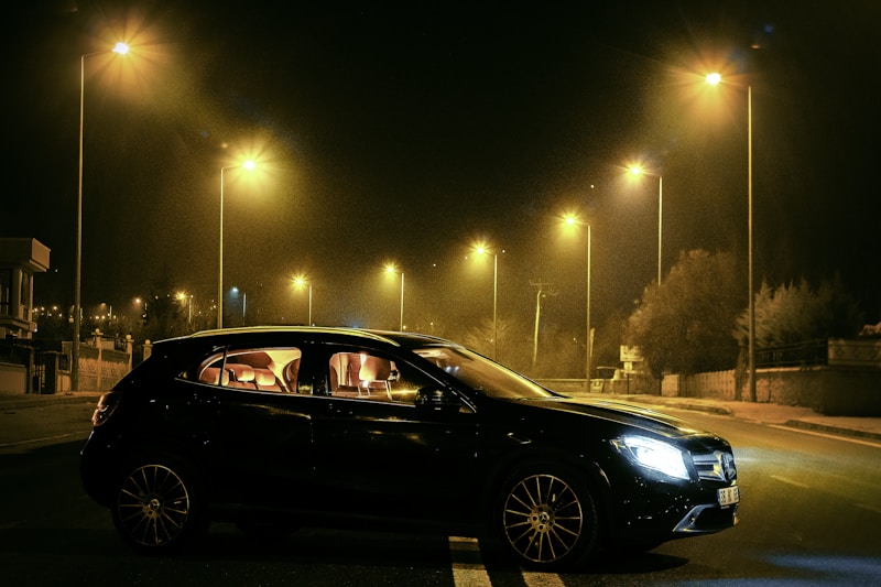 A sleek black car is parked under multiple streetlights on a dimly lit road during nighttime. The lights of the lamps cast a warm yellow glow, creating contrasting shadows around the vehicle. The interior of the car is softly illuminated, highlighting its plush seats. Sparse trees and some buildings are visible in the background.