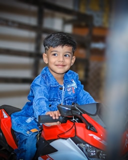 A young child with a happy expression is sitting on a toy motorcycle. The child is dressed in a denim jacket and appears to be enjoying themselves.