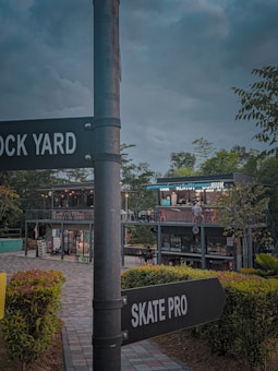 A two-story modern structure made from shipping containers serves as a café or restaurant in an outdoor area. Surrounding the structure is a landscaped environment with various plants and bushes. The sky is overcast, creating a subdued atmosphere. A prominent signpost in the foreground displays directions to 'Rock Yard' and 'Skate Pro'.