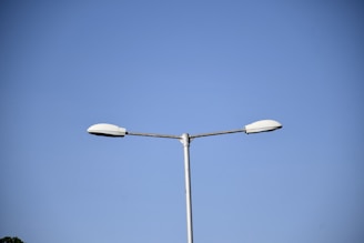 A single streetlight with two lamps extends against a clear blue sky. The streetlight is centered in the image, and the lamps are simple and modern in design. The surrounding sky is cloudless, enhancing the stark contrast between the grayish metal of the streetlight and the blue background.