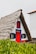 A traditional, triangular-shaped house with a distinctive thatched roof and vibrant red door and window trim. The facade is white, accented with blue and black shutters, surrounded by lush green foliage in the foreground.