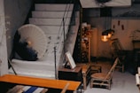 A cozy, rustic interior featuring a set of stairs adorned with a large parasol and ink brush art. The room contains bamboo furniture including a chair and small table. Warm lighting from hanging lamps creates a comfortable ambiance, complemented by additional decorative elements like traditional calligraphy scrolls.