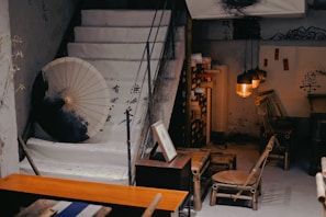A cozy, rustic interior featuring a set of stairs adorned with a large parasol and ink brush art. The room contains bamboo furniture including a chair and small table. Warm lighting from hanging lamps creates a comfortable ambiance, complemented by additional decorative elements like traditional calligraphy scrolls.