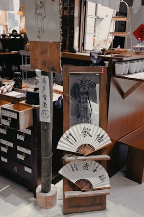 The interior space resembles a traditional Asian shop or tea house, displaying various items like wooden drawers with labels, a bamboo pole, traditional fans with calligraphy, and other cultural artifacts. There is a drawing of a figure on a piece of paper and shelves containing pottery and tea sets. The setting is warm, inviting, and full of vintage charm.