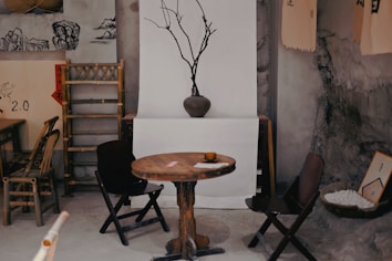 A rustic interior setting with a wooden table and chairs. A bare branch in a vase serves as a minimalist centerpiece. The decor includes bamboo furniture, traditional art prints, and a bowl filled with small white stones. The walls have a rough, earthy texture, and there are various traditional decorations such as scrolls hanging on a backdrop.