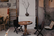 A rustic interior setting with a wooden table and chairs. A bare branch in a vase serves as a minimalist centerpiece. The decor includes bamboo furniture, traditional art prints, and a bowl filled with small white stones. The walls have a rough, earthy texture, and there are various traditional decorations such as scrolls hanging on a backdrop.