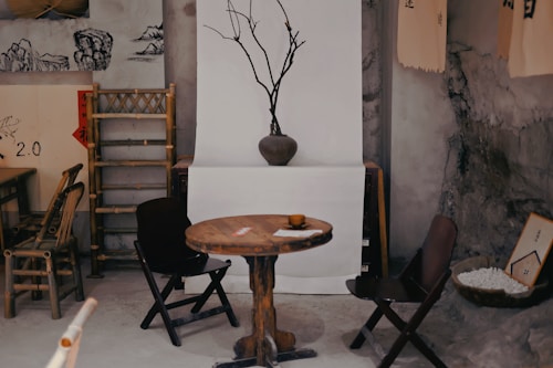 A rustic interior setting with a wooden table and chairs. A bare branch in a vase serves as a minimalist centerpiece. The decor includes bamboo furniture, traditional art prints, and a bowl filled with small white stones. The walls have a rough, earthy texture, and there are various traditional decorations such as scrolls hanging on a backdrop.