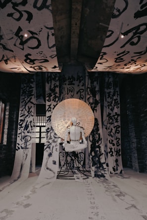 An artistic installation features a white humanoid figure seated in a meditative pose, surrounded by large sheets of calligraphy hanging from the ceiling and walls. The sheets contain bold, black brush strokes of Asian characters. A circular backdrop with additional writing illuminates the figure, casting an ethereal glow.