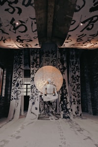An artistic installation features a white humanoid figure seated in a meditative pose, surrounded by large sheets of calligraphy hanging from the ceiling and walls. The sheets contain bold, black brush strokes of Asian characters. A circular backdrop with additional writing illuminates the figure, casting an ethereal glow.