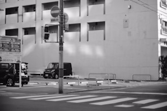 A black and white urban scene depicting a parking lot in front of a multi-story building. There is a traffic light and some parked vehicles, including two cars visible. A sign with rates for parking can be seen, and the architecture includes balconies and rectangular shapes.