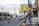 A city street lined with buildings, decorated lamp posts, and banners, with traffic cones dividing the lanes. Various vehicles, including cars and motorcycles, are present. A large sign in Indonesian advertising real estate is prominently displayed.