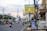 A city street lined with buildings, decorated lamp posts, and banners, with traffic cones dividing the lanes. Various vehicles, including cars and motorcycles, are present. A large sign in Indonesian advertising real estate is prominently displayed.