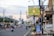 A city street lined with buildings, decorated lamp posts, and banners, with traffic cones dividing the lanes. Various vehicles, including cars and motorcycles, are present. A large sign in Indonesian advertising real estate is prominently displayed.
