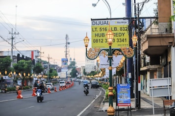 A city street lined with buildings, decorated lamp posts, and banners, with traffic cones dividing the lanes. Various vehicles, including cars and motorcycles, are present. A large sign in Indonesian advertising real estate is prominently displayed.