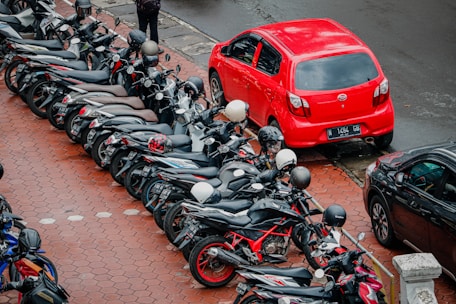 A lineup of motorcycles parked closely alongside a brightly colored red car on a paved, hexagonal patterned sidewalk. Most motorcycles are black, with several helmets resting on the seats. The background includes sections of another black car and the edge of the street.