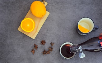 An overhead view of a kitchen scene featuring a yellow passion fruit and a half orange placed on a light wooden cutting board. Nearby, a handful of star anise is scattered across the surface. A hand is pouring a dark liquid from a glass bottle into a cup, which is positioned next to another cup containing an orange slice.