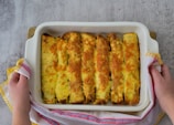A pair of hands holds a rectangular white baking dish containing six rolled, baked items topped with melted cheese, resting on a light wooden surface. A colorful kitchen towel with pink and yellow stripes is wrapped around the dish.