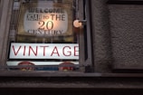 A vintage shop window displays a sign that reads 'Welcome Back to the 20th Century' with large, prominent letters spelling 'VINTAGE' beneath it. The sign is housed in a wooden frame, and the letters are illuminated against a textured wall.