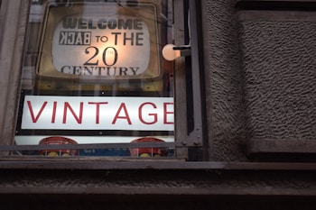A vintage shop window displays a sign that reads 'Welcome Back to the 20th Century' with large, prominent letters spelling 'VINTAGE' beneath it. The sign is housed in a wooden frame, and the letters are illuminated against a textured wall.