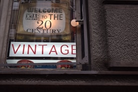 A vintage shop window displays a sign that reads 'Welcome Back to the 20th Century' with large, prominent letters spelling 'VINTAGE' beneath it. The sign is housed in a wooden frame, and the letters are illuminated against a textured wall.