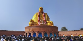 A large golden statue sits in a meditative pose on a circular platform adorned with intricate carvings. The statue is draped with a garland and surrounded by a crowd of people. The sky is clear and blue, enhancing the vibrant colors of the scene. The platform features arches and decorative details.
