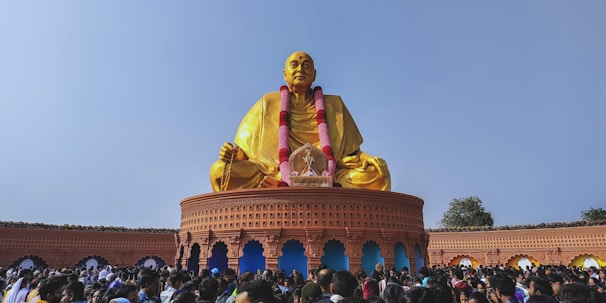 A large golden statue sits in a meditative pose on a circular platform adorned with intricate carvings. The statue is draped with a garland and surrounded by a crowd of people. The sky is clear and blue, enhancing the vibrant colors of the scene. The platform features arches and decorative details.