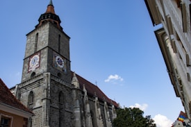 A tall, historic stone church tower with a clock face on its side stands against a clear blue sky. The architecture features detailed stonework with a pointed roof and small crosses at the apex. Adjacent buildings with pitched roofs and a tree are visible, with part of the Romanian flag seen on one building.