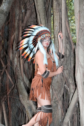 A person in traditional attire with a feathered headdress stands against a backdrop of large tree roots. The outfit includes a brown fringed garment and ornamental accessories. The setting appears to be a forest with dense wooden textures and hints of green foliage in the background.
