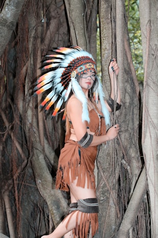 A person in traditional attire with a feathered headdress stands against a backdrop of large tree roots. The outfit includes a brown fringed garment and ornamental accessories. The setting appears to be a forest with dense wooden textures and hints of green foliage in the background.