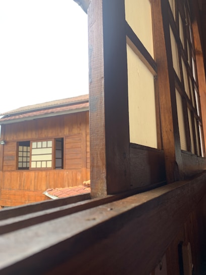 A view through a wooden window frame of a traditional wooden building. The window and building have wooden panels with a warm, natural finish. The outer building features a similar wooden texture with window panels divided by grids, highlighting a classic architectural style.