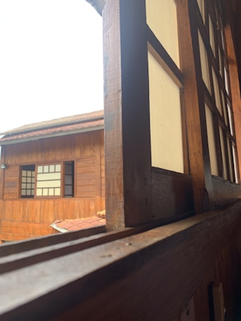A view through a wooden window frame of a traditional wooden building. The window and building have wooden panels with a warm, natural finish. The outer building features a similar wooden texture with window panels divided by grids, highlighting a classic architectural style.