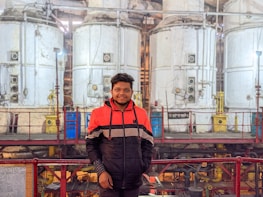 A person stands in front of large, industrial machinery with several cylindrical tanks. The machinery has a utilitarian design with visible pipes and valves. The person is wearing a red and black jacket, standing on a platform with railings.