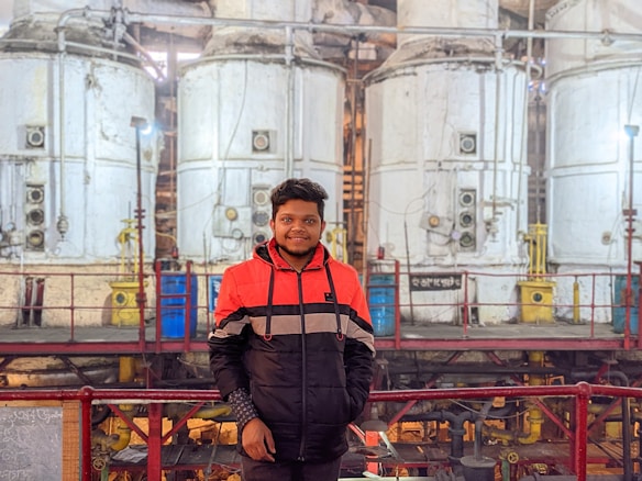 A person stands in front of large, industrial machinery with several cylindrical tanks. The machinery has a utilitarian design with visible pipes and valves. The person is wearing a red and black jacket, standing on a platform with railings.