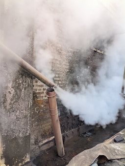 A rusty metal pipe is emitting a thick cloud of white smoke against a weathered brick wall. The environment appears industrial and gritty, with visible grime on the ground and walls.
