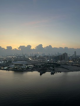 The image captures a panoramic view of a cityscape at dawn or dusk. The skyline is lined with various high-rise buildings, some of which are illuminated by artificial lights. The foreground features a calm body of water reflecting the dim ambient light. Above, a vast expanse of sky transitions smoothly from lighter to darker hues, marked by thick, fluffy clouds just above the horizon.