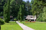 A quaint house with a wooden roof nestled in a lush green garden. Tall trees surround the area, and a pathway leads to the house. There are outdoor tables with umbrellas and a small circular garden with red flowers.