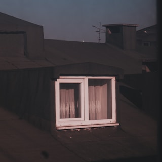 A rooftop with a small white-framed window, partially covered by sheer curtains that allow some light to pass through. Surrounding the window are dark shingles or roofing material. An antenna is visible in the background against a slightly hazy sky.
