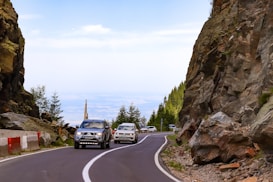 Two cars are driving along a winding mountain road, bordered by large rocky cliffs on one side and a mix of greenery on the other. The scene conveys a sense of movement and adventure as the vehicles navigate the curving path. The sky is bright and clear, adding to the expansive feeling of the landscape.