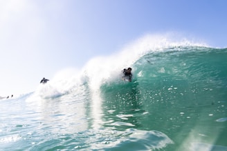 A surfer rides a powerful turquoise wave with skill and balance, surrounded by splashes of water. The sun is shining brightly in a clear blue sky, and another surfer can be seen in the background, navigating the wave. The water is crystal clear, showcasing the dynamic motion and energy of the surf.