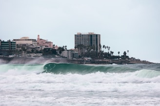 Large ocean waves crash near the shoreline with a cityscape in the background. The skyline features tall buildings and palm trees lining the coast, suggesting a coastal urban environment.
