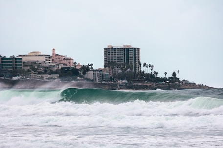 Large ocean waves crash near the shoreline with a cityscape in the background. The skyline features tall buildings and palm trees lining the coast, suggesting a coastal urban environment.